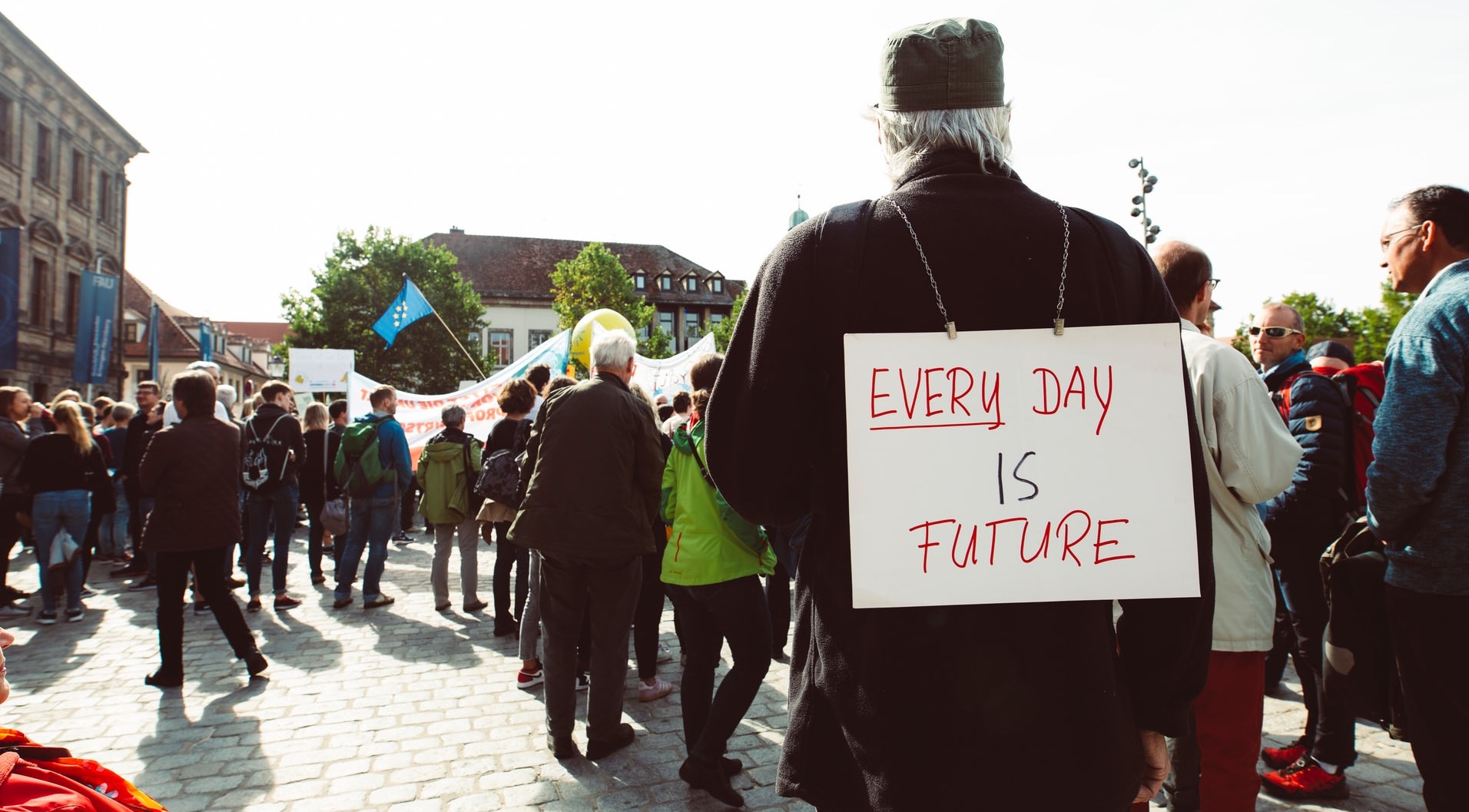 man stands towards a crowd with a sign on his back that reads 'every day is future'